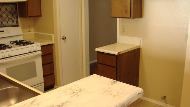 kitchen featuring baseboards, white range with gas cooktop, light countertops, tile patterned floors, and brown cabinets