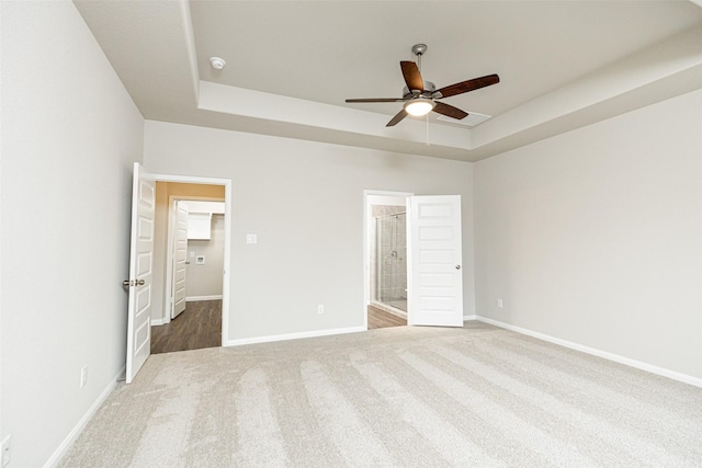 unfurnished bedroom featuring a ceiling fan, ensuite bathroom, a tray ceiling, carpet floors, and baseboards