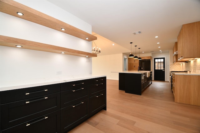 kitchen with a center island with sink, black fridge, dark cabinetry, light wood finished floors, and decorative backsplash