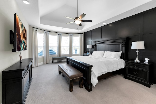 bedroom with a decorative wall, light colored carpet, a raised ceiling, and a ceiling fan