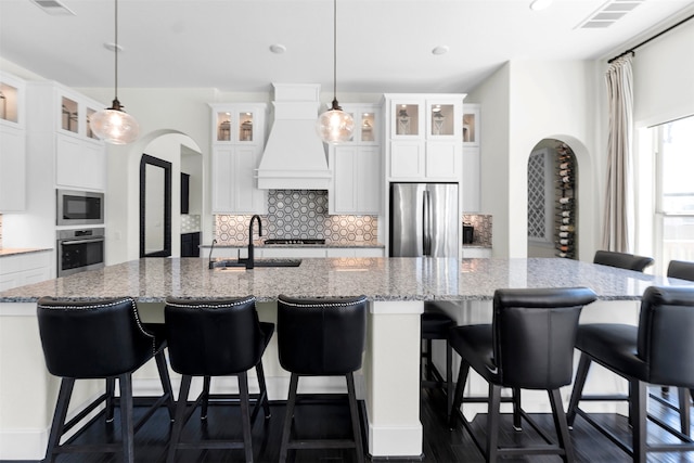 kitchen featuring visible vents, arched walkways, a large island with sink, stainless steel appliances, and custom range hood