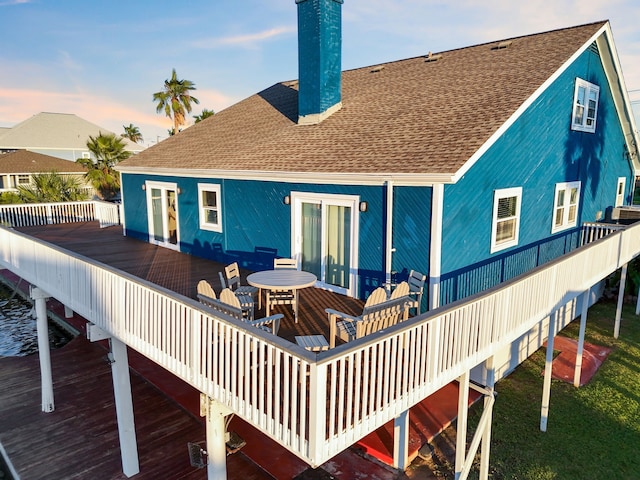 back of house with a deck, outdoor dining space, a chimney, and a shingled roof