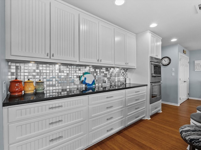 kitchen featuring double oven, dark wood-style flooring, decorative backsplash, white cabinets, and dark countertops