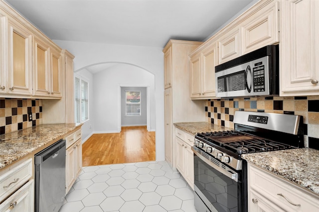 kitchen featuring light stone counters, decorative backsplash, appliances with stainless steel finishes, light tile patterned flooring, and arched walkways