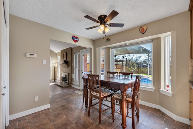 dining space featuring baseboards, a textured ceiling, a brick fireplace, and ceiling fan