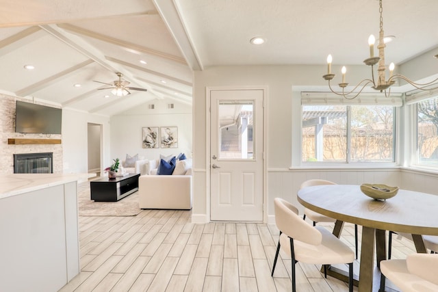 dining area with light wood finished floors, vaulted ceiling with beams, a wainscoted wall, a stone fireplace, and ceiling fan with notable chandelier