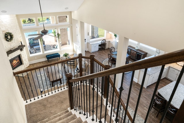 stairs featuring wood finished floors, a towering ceiling, a stone fireplace, baseboards, and ceiling fan