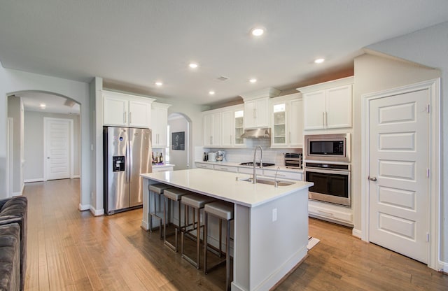 kitchen with arched walkways, appliances with stainless steel finishes, under cabinet range hood, and a kitchen island with sink