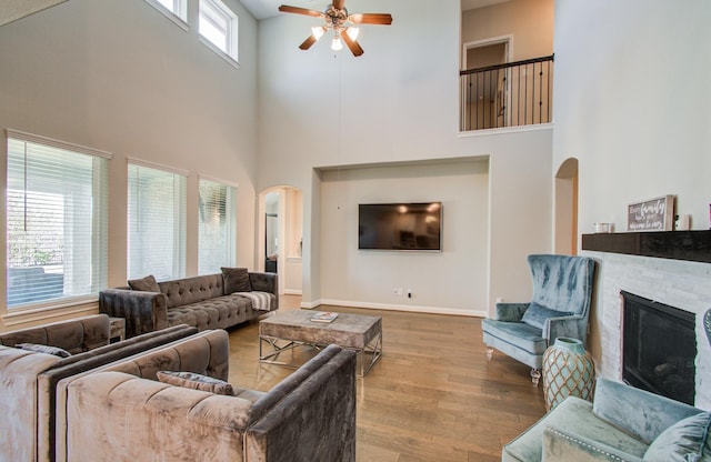 living area featuring wood finished floors, arched walkways, a stone fireplace, baseboards, and ceiling fan