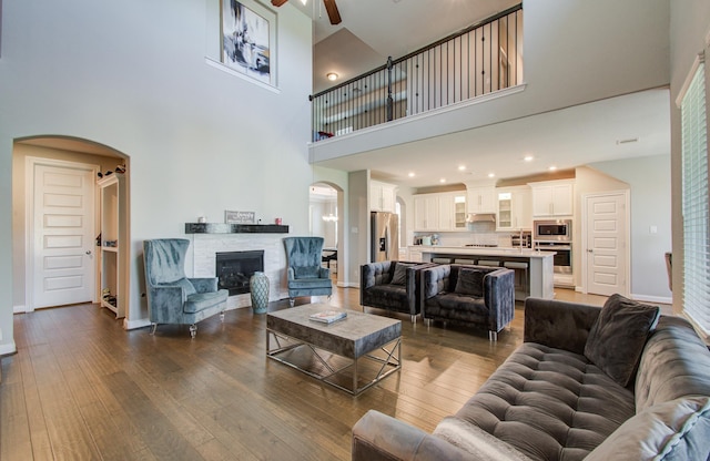 living room with arched walkways, a stone fireplace, dark wood-type flooring, and ceiling fan