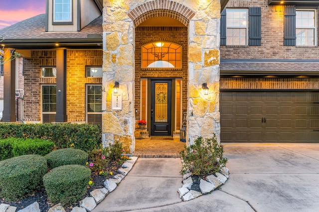 exterior entry at dusk with stone siding, roof with shingles, concrete driveway, an attached garage, and brick siding