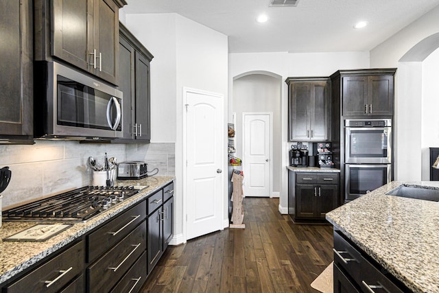 kitchen with dark wood finished floors, light stone counters, decorative backsplash, appliances with stainless steel finishes, and arched walkways