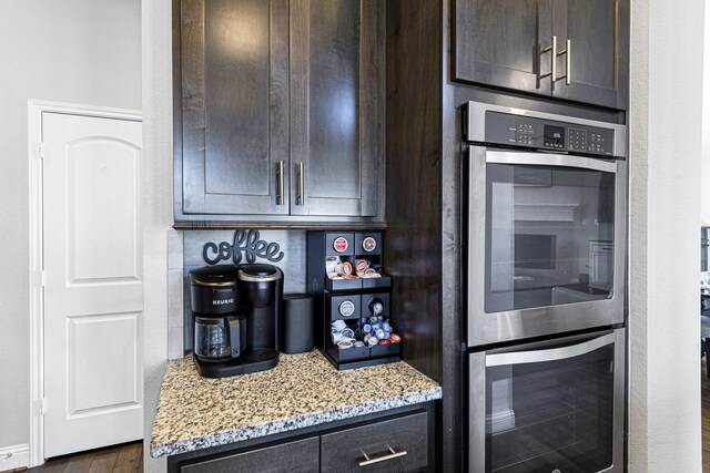 kitchen with stainless steel double oven, light stone countertops, dark brown cabinetry, and dark wood-style floors