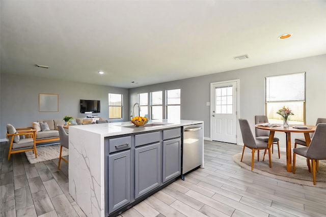 kitchen featuring wood finish floors, a kitchen island with sink, gray cabinets, a sink, and dishwasher