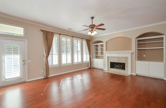 unfurnished living room with built in shelves, wood finished floors, crown molding, and a tiled fireplace