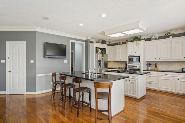 kitchen with dark countertops, decorative backsplash, appliances with stainless steel finishes, wood finished floors, and a kitchen island with sink