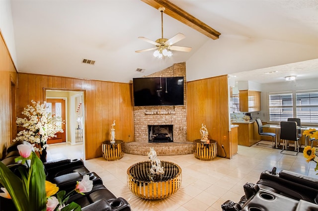 living area featuring wooden walls, visible vents, a brick fireplace, beam ceiling, and a ceiling fan