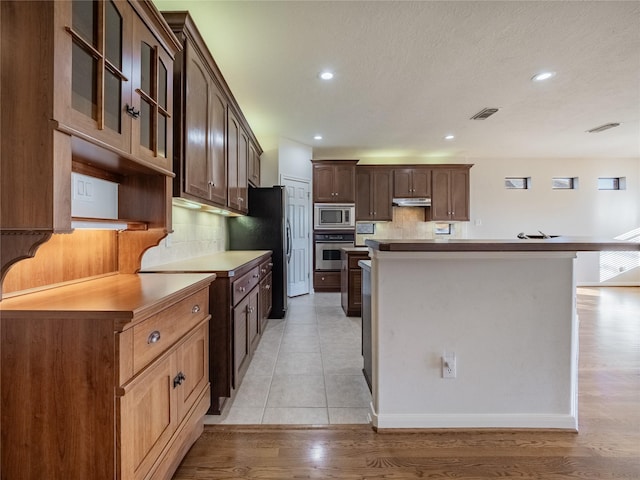 kitchen featuring tasteful backsplash, dark brown cabinets, glass insert cabinets, light wood-style flooring, and appliances with stainless steel finishes