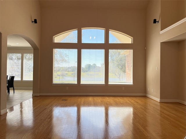 spare room featuring arched walkways, light wood-style flooring, a high ceiling, and baseboards