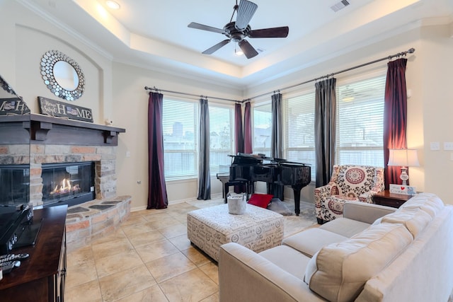 living room with a ceiling fan, visible vents, a tray ceiling, a stone fireplace, and crown molding