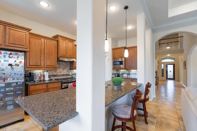 kitchen featuring dark stone counters, arched walkways, stainless steel appliances, under cabinet range hood, and a kitchen breakfast bar