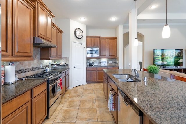 kitchen featuring dark stone countertops, brown cabinetry, a sink, stainless steel appliances, and under cabinet range hood