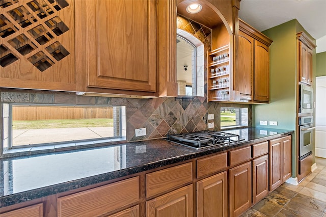 kitchen with brown cabinets, dark stone counters, appliances with stainless steel finishes, stone finish flooring, and tasteful backsplash