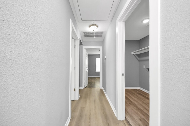 corridor with visible vents, light wood finished floors, baseboards, attic access, and a textured wall