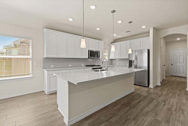 kitchen with an island with sink, arched walkways, a sink, stainless steel appliances, and white cabinetry