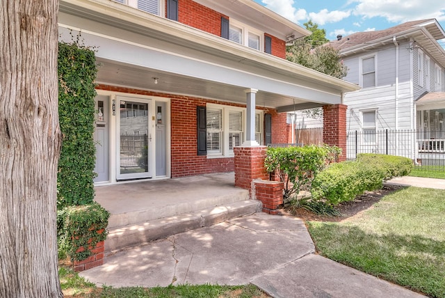 doorway to property featuring brick siding, covered porch, and fence