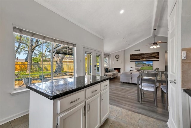 kitchen featuring dark stone countertops, light tile patterned floors, a warm lit fireplace, vaulted ceiling, and white cabinets