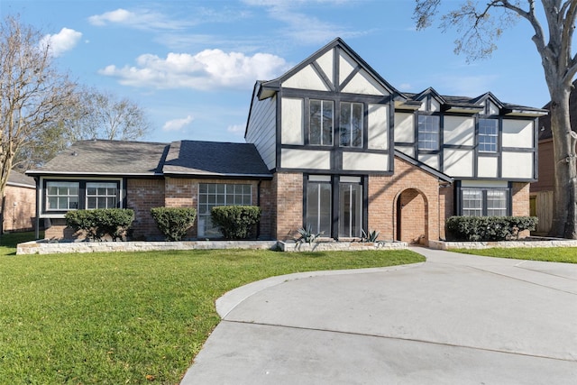 tudor-style house with driveway, roof with shingles, stucco siding, a front lawn, and brick siding