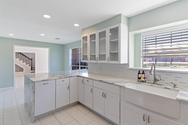 kitchen featuring light stone counters, a peninsula, a sink, white cabinets, and glass insert cabinets