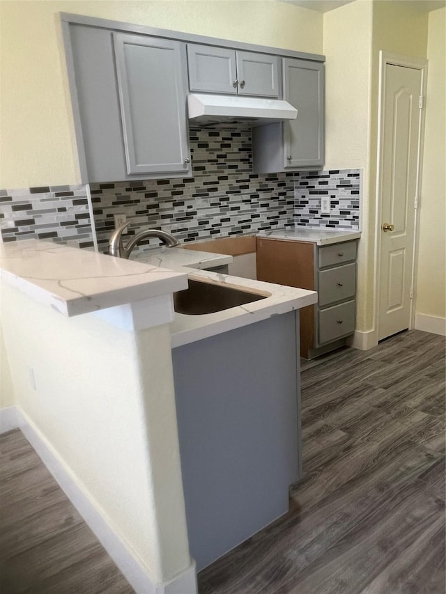 kitchen with dark wood-style flooring, gray cabinets, a sink, decorative backsplash, and under cabinet range hood