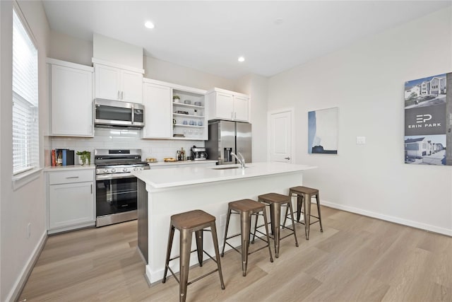 kitchen featuring open shelves, a kitchen island with sink, decorative backsplash, light wood-style floors, and appliances with stainless steel finishes