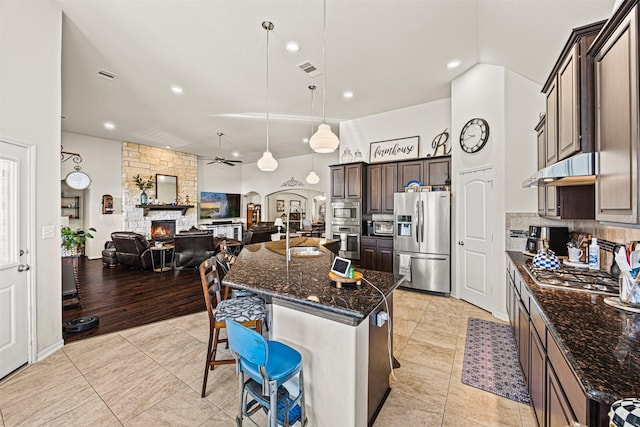 kitchen featuring a fireplace, ceiling fan, stainless steel appliances, dark brown cabinets, and under cabinet range hood