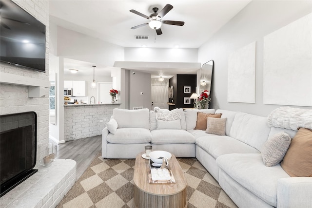 living area with ceiling fan, a brick fireplace, visible vents, and light wood-type flooring