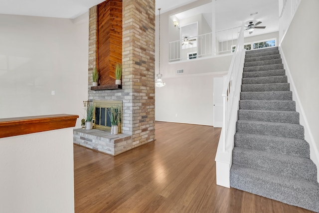 stairs with visible vents, wood finished floors, a high ceiling, a brick fireplace, and ceiling fan