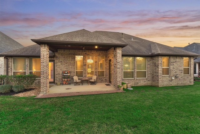 back of house at dusk featuring brick siding, a lawn, a patio, and roof with shingles