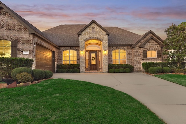 french country inspired facade featuring driveway, a front yard, a shingled roof, a garage, and brick siding