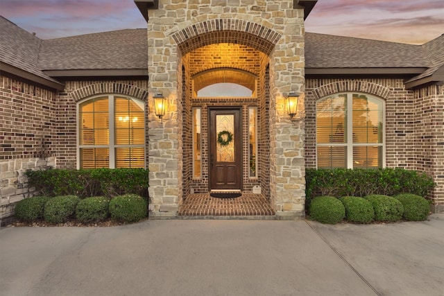 exterior entry at dusk featuring brick siding, stone siding, and a shingled roof