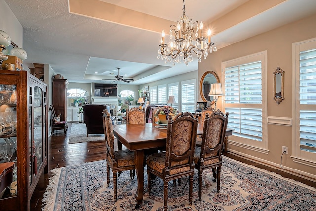 dining room with baseboards, dark wood finished floors, a tray ceiling, a fireplace, and ceiling fan with notable chandelier