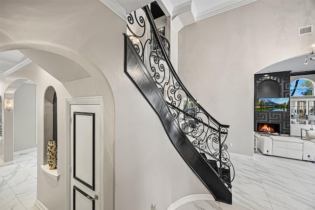 staircase featuring visible vents, a warm lit fireplace, marble finish floor, and crown molding