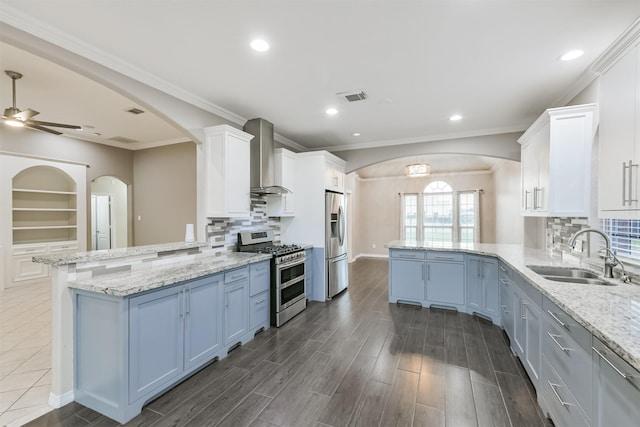 kitchen featuring wall chimney range hood, appliances with stainless steel finishes, a peninsula, arched walkways, and a sink