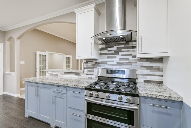 kitchen featuring wall chimney range hood, light stone counters, arched walkways, stainless steel gas stove, and white cabinets