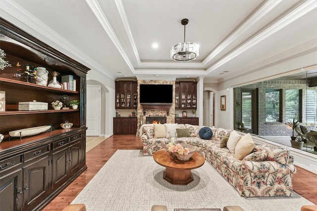 living area with light wood finished floors, arched walkways, ornamental molding, a stone fireplace, and a raised ceiling