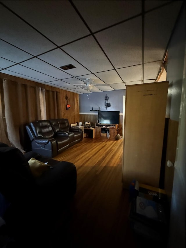 living area with a ceiling fan, wood finished floors, visible vents, a drop ceiling, and wood walls