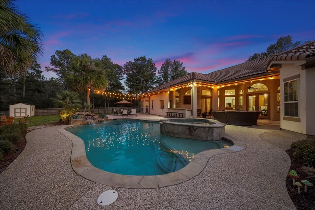 pool at dusk featuring a pool with connected hot tub, a shed, a fenced backyard, an outdoor structure, and a patio