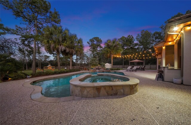 pool at dusk with an outbuilding, a pool with connected hot tub, a storage shed, and a patio