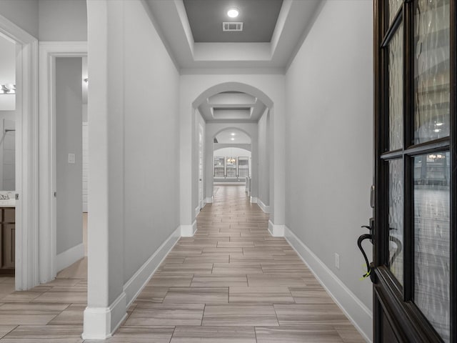 hall featuring baseboards, visible vents, wood tiled floor, arched walkways, and a raised ceiling